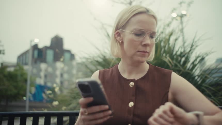 A stylish blonde woman wearing glasses sitting on a city bench smiles happily after checking her smartwatch