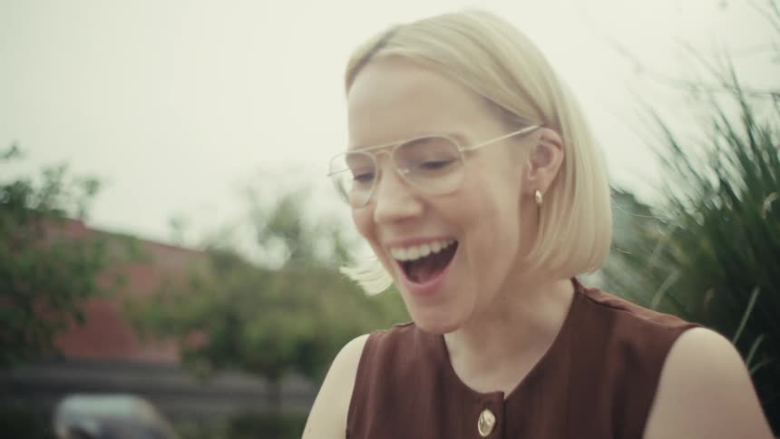 A stylish blonde woman sitting on a park bench in the city reacts with surprise and joy to something she sees on her smartphone