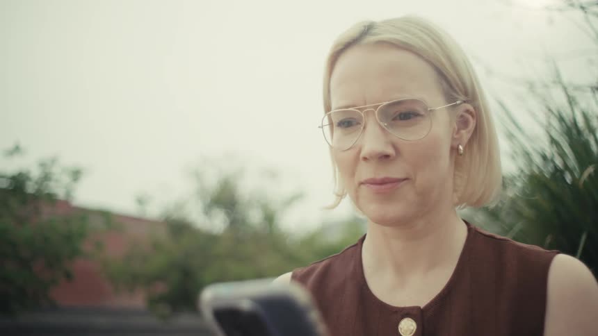 A stylish blonde woman with glasses pauses from her phone, looking up with a gentle, hopeful smile while sitting on a city park bench