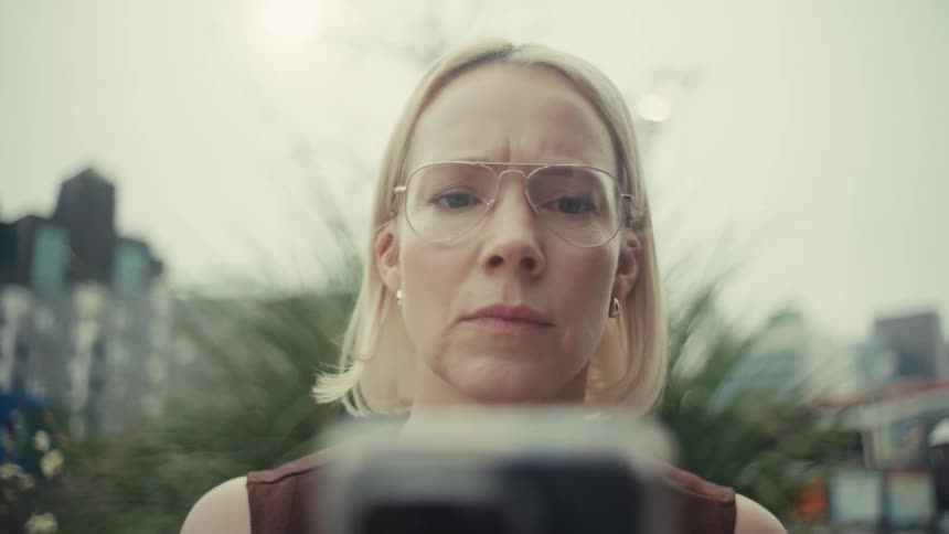 A concerned blonde woman in glasses focuses intently on her smartphone screen while outdoors in the city