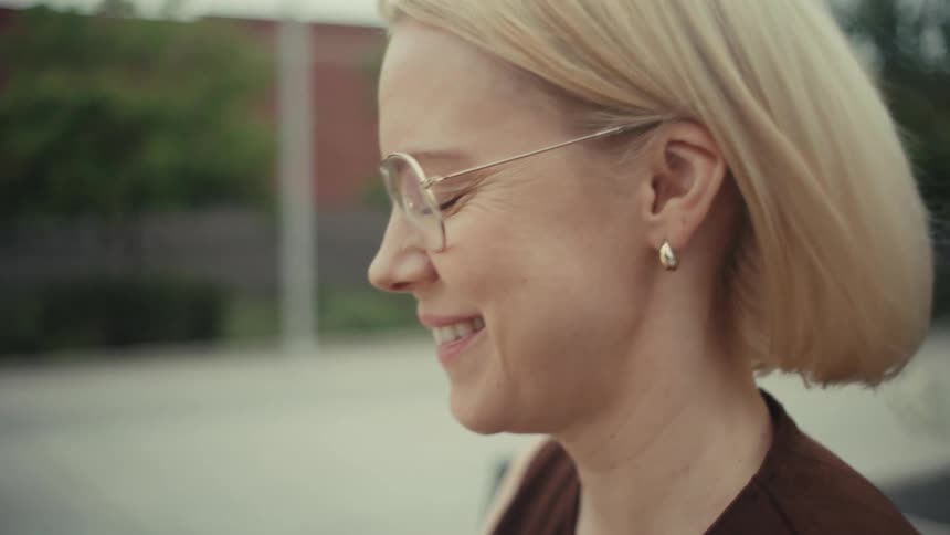 A smiling blonde woman with glasses uses her smartphone while sitting outside on a city bench