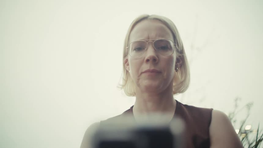 A serious blonde woman with glasses intently reads something on her phone, shot from a low angle