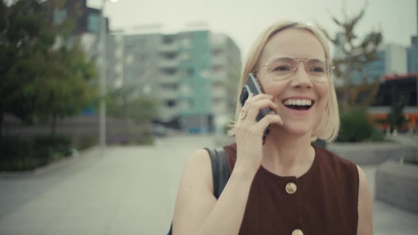 A cheerful blonde businesswoman in glasses laughs and talks on her smartphone while walking through a modern city park in slow motion