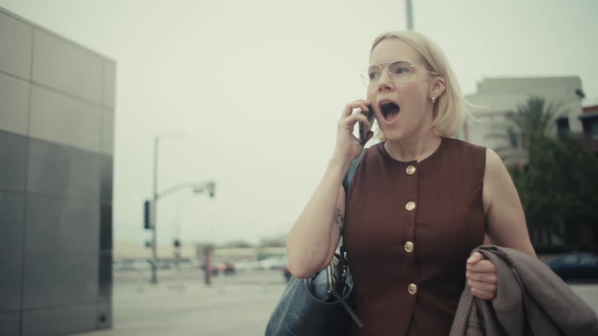A blonde businesswoman in glasses has an expressive and emotional phone conversation while walking on a city street in slow motion