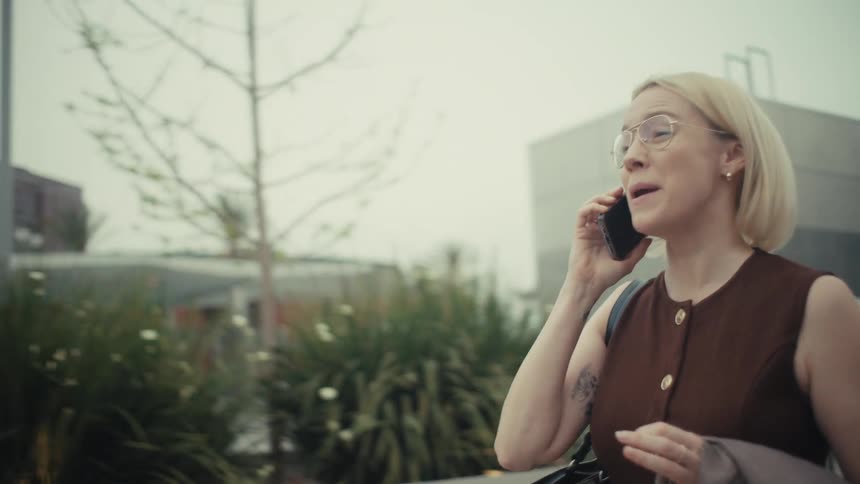 Slow motion of a professional blonde woman having a happy phone call that turns into a serious conversation as she walks through the city