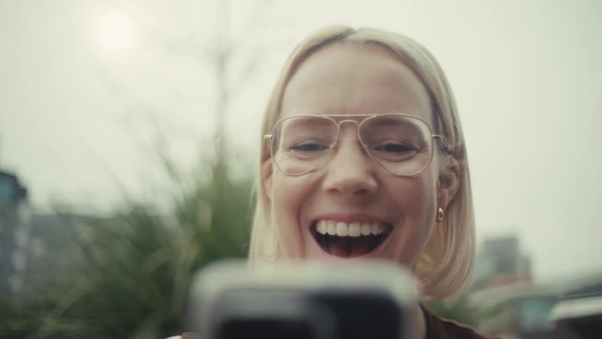 A stylish blonde woman's expression transforms from focused concentration to surprised delight and laughter as she looks at a screen outdoors