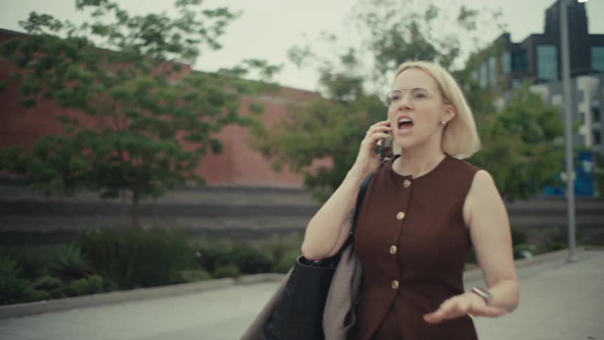 A stylish woman walks down a city street, answers her phone, and erupts in ecstatic celebration, pumping her fist in victory