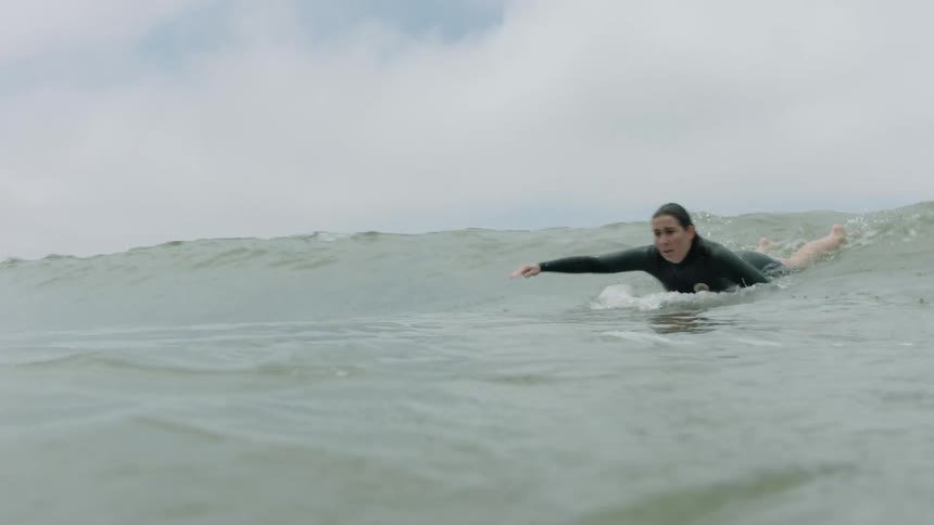 A female surfer in a black wetsuit paddles intensely to catch a wave