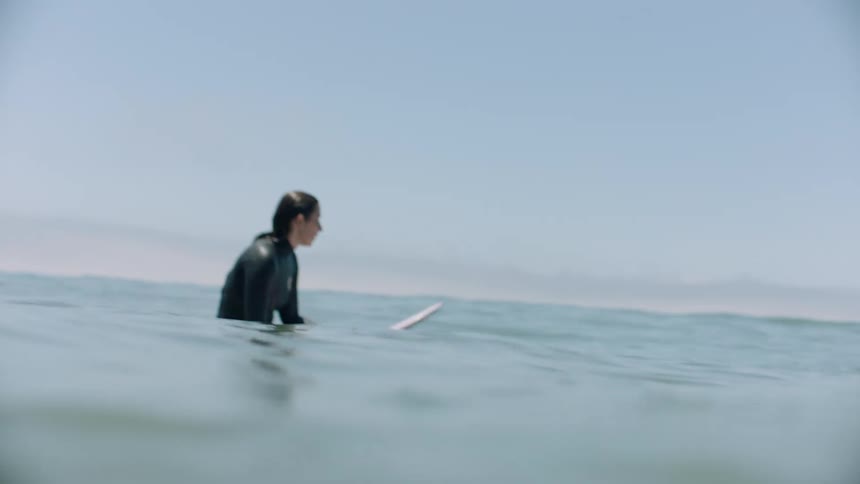 A female surfer in a wetsuit patiently sits on her board in the gentle ocean swells, waiting for the perfect wave