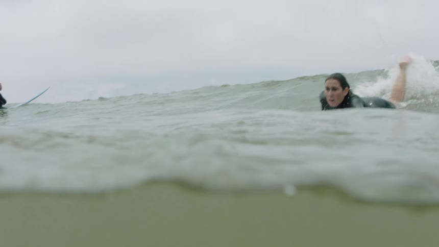 A determined female surfer in a wetsuit paddles hard to catch a wave, filmed in normal speed from the water's surface