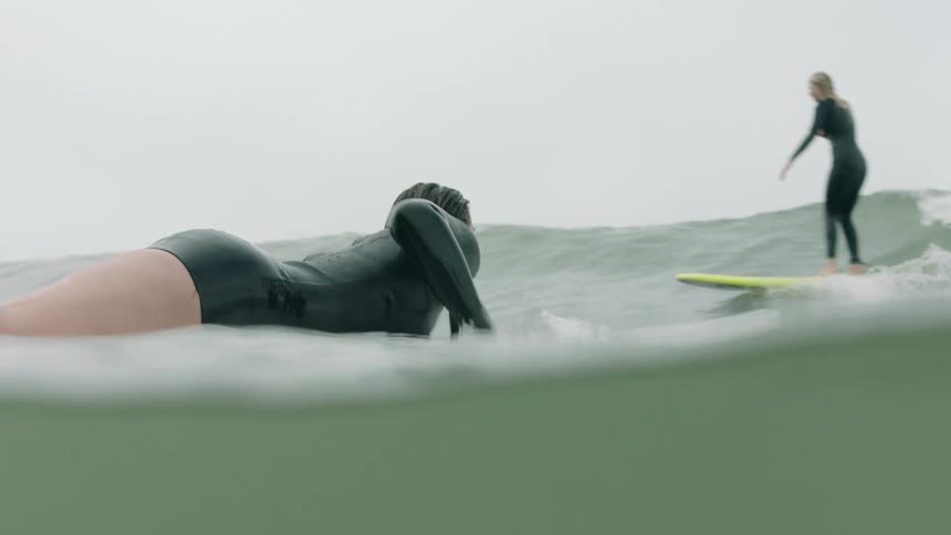 From an immersive water-level view, a female surfer in a wetsuit ducks under a crashing ocean wave in slow motion