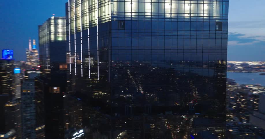 An elegant aerial shot pulls back from a modern glass skyscraper, revealing the vibrant New York City skyline at dusk