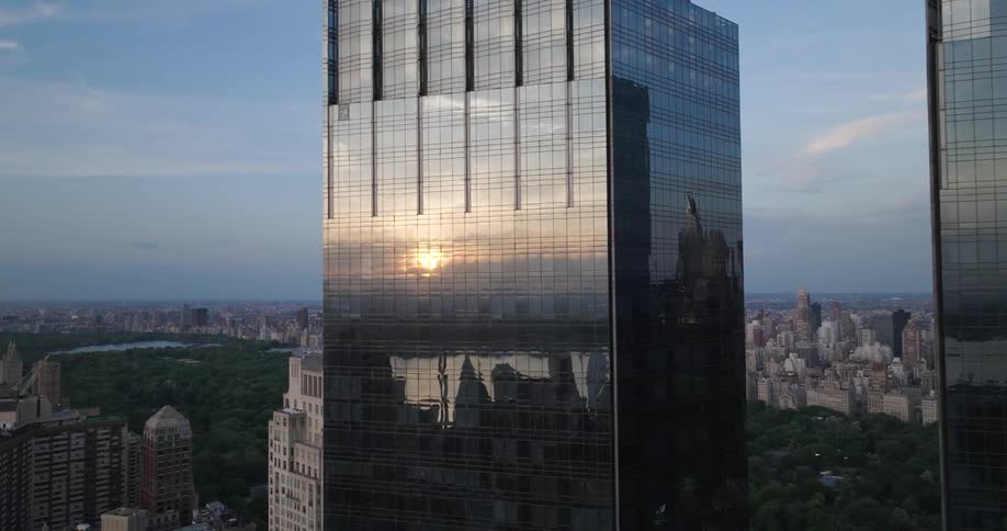 An aerial view slowly pushes in on a modern glass skyscraper reflecting the golden sunset over the Manhattan skyline and Central Park
