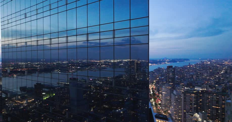 An aerial pullback reveals the stunning New York City skyline at dusk, with the vibrant city lights reflected in the glass windows of a modern skyscraper