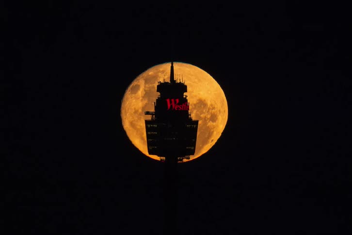 A time-lapse of a brilliant orange supermoon rising and passing behind the silhouetted Sydney Tower Eye at night