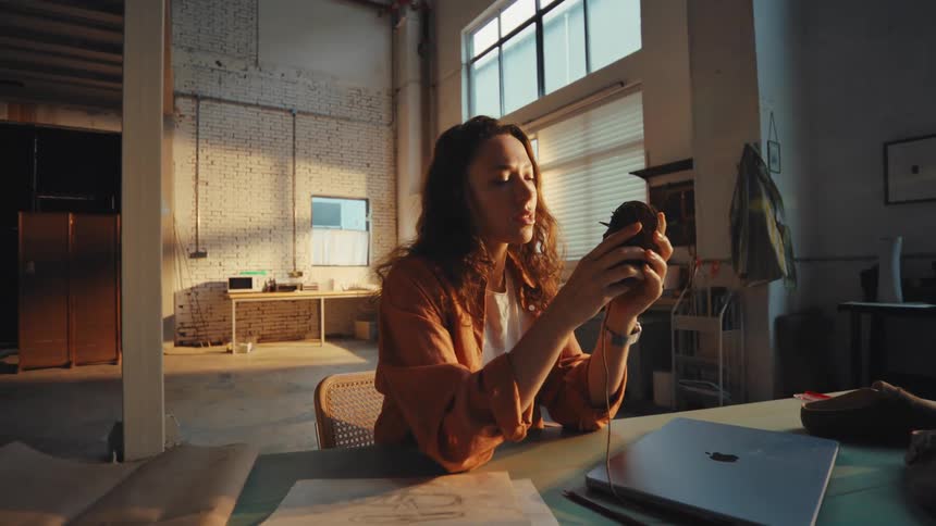 A thoughtful young woman takes a break to drink from a mug while sitting at her desk in a sunlit industrial art studio