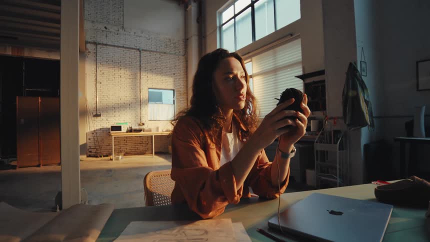 A thoughtful young woman takes a break to drink from a mug while sitting at her desk in a sunlit industrial art studio