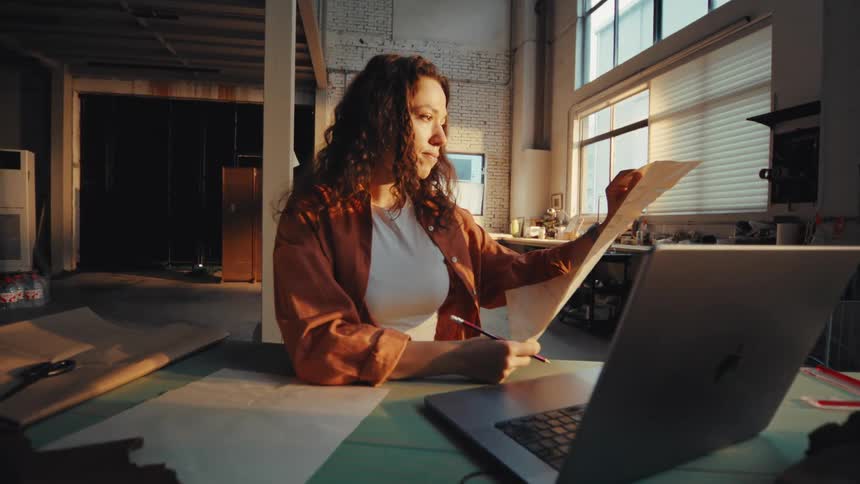 A focused female designer reviews a paper pattern at her workbench while working on her laptop in a sunlit industrial loft studio