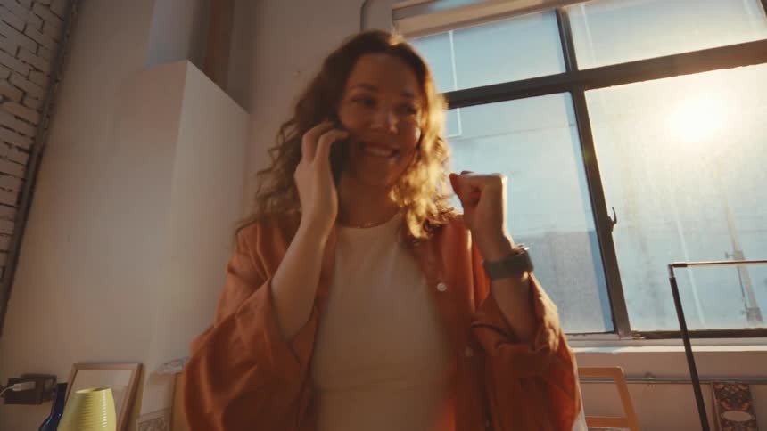 Smiling woman receives exciting news on a phone call and celebrates with a fist pump in her sun-drenched studio apartment