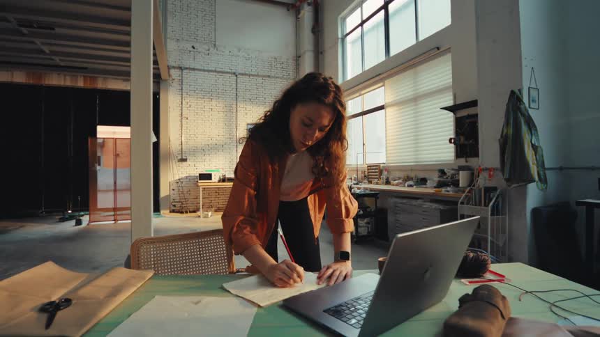 A focused young female artisan sketches a design on paper at her workbench in a sunlit industrial workshop