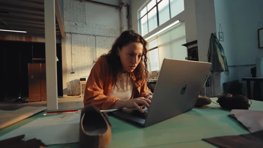 A diligent female artisan works on a new shoe design in her workshop, using a laptop for research and sketching ideas in her notebook