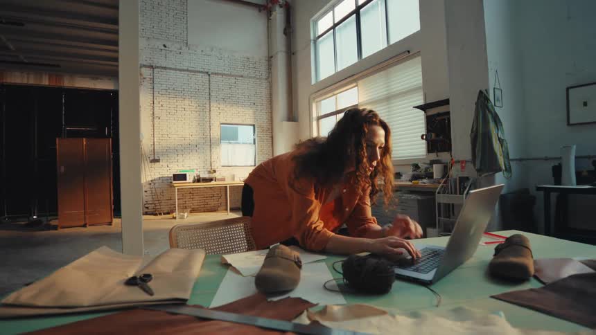A focused female shoe designer works diligently on her laptop in a sunlit industrial workshop
