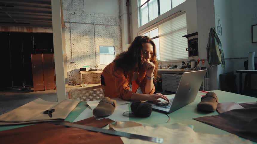 A focused female shoe designer works diligently on her laptop in a sunlit industrial workshop