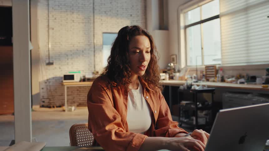 A focused woman finishes her work, closes her laptop with a smile, and relaxes in a sunlit loft-style studio