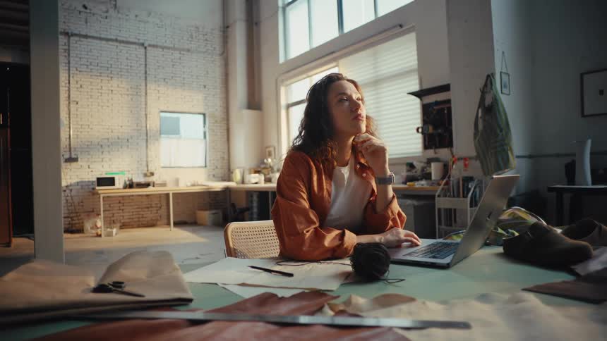 A thoughtful female artisan contemplates her next project while working on her laptop in a spacious, sunlit workshop