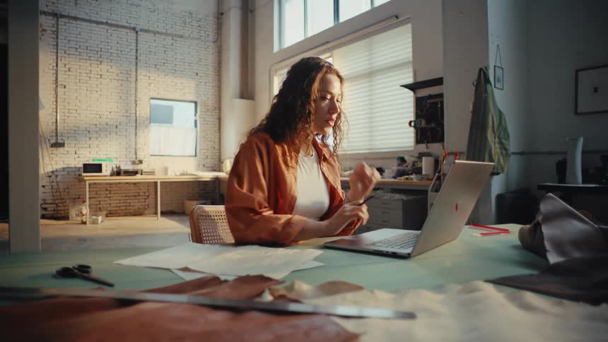 A thoughtful young female artisan works on her laptop in a spacious, sunlit creative studio