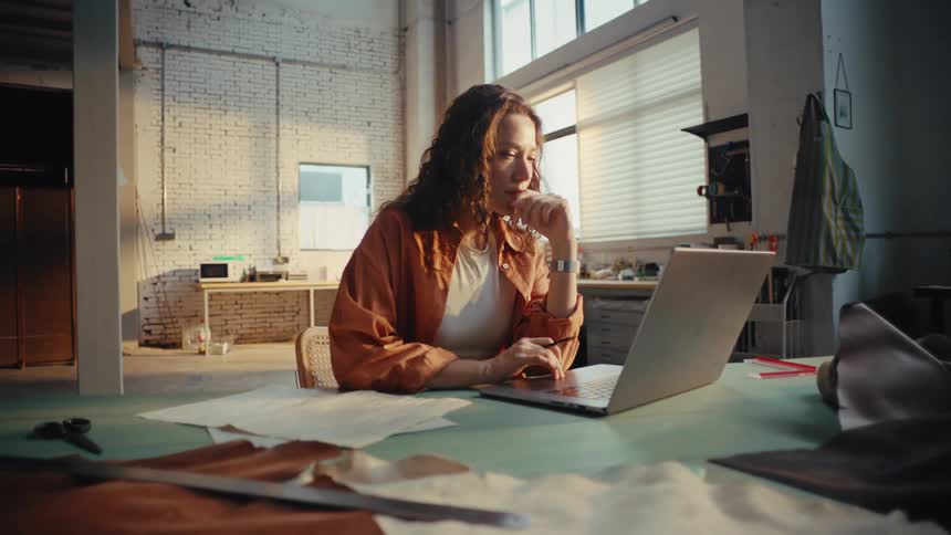 A thoughtful young female artisan works on her laptop in a spacious, sunlit creative studio