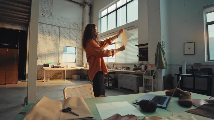 A creative female artisan inspects a paper design pattern in her spacious, sunlit industrial loft studio