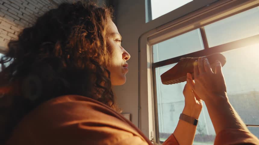 A female shoemaker with curly hair carefully inspects a handmade shoe in her sunlit workshop