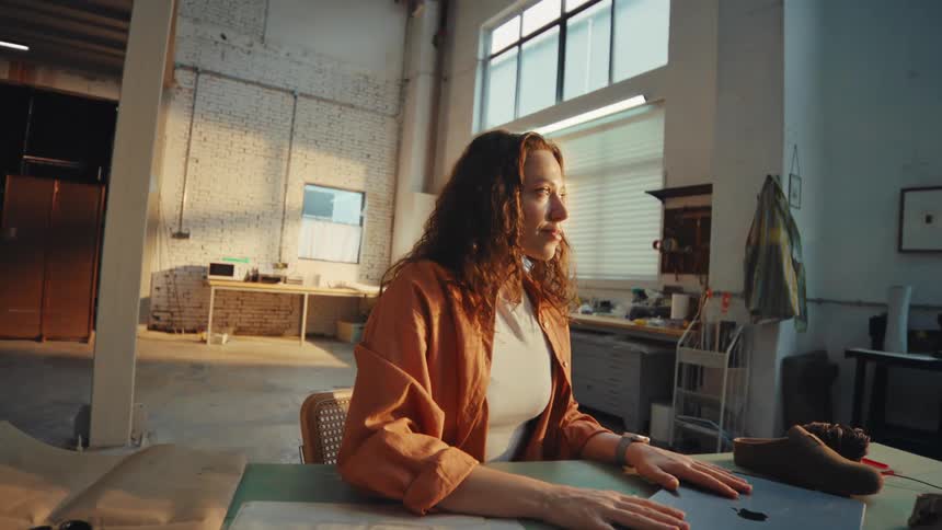 A female designer finishes typing on her laptop, closes it with a smile, and takes a moment of satisfaction in her spacious, sunlit workshop