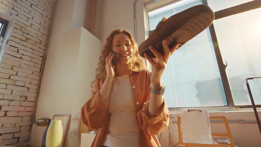 A cheerful female entrepreneur in her sunlit workshop holds up a suede shoe while confirming details over a phone call