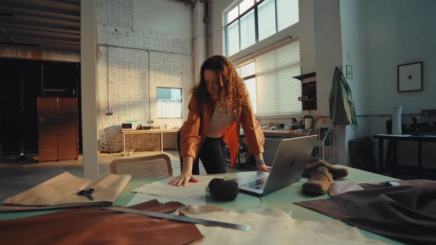 A female artisan works on her laptop, surrounded by leather materials and patterns in a spacious industrial workshop