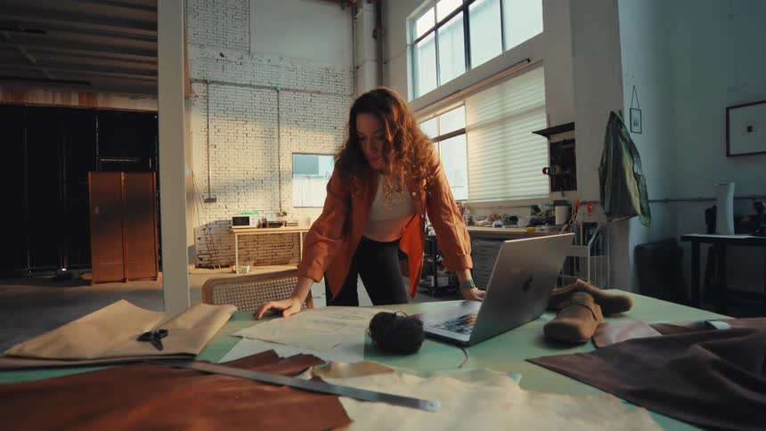 A female artisan works on her laptop, surrounded by leather materials and patterns in a spacious industrial workshop