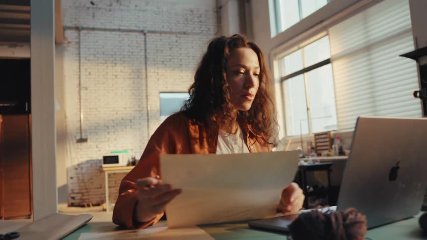 A focused female designer references her sketches while working on a laptop in her sun-drenched workshop