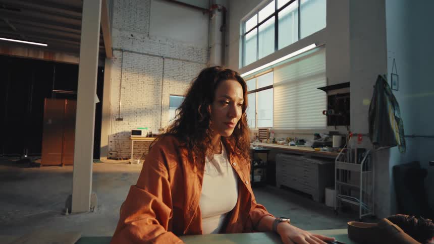 A young female artisan closes her laptop after finishing work for the day in her sunlit industrial studio