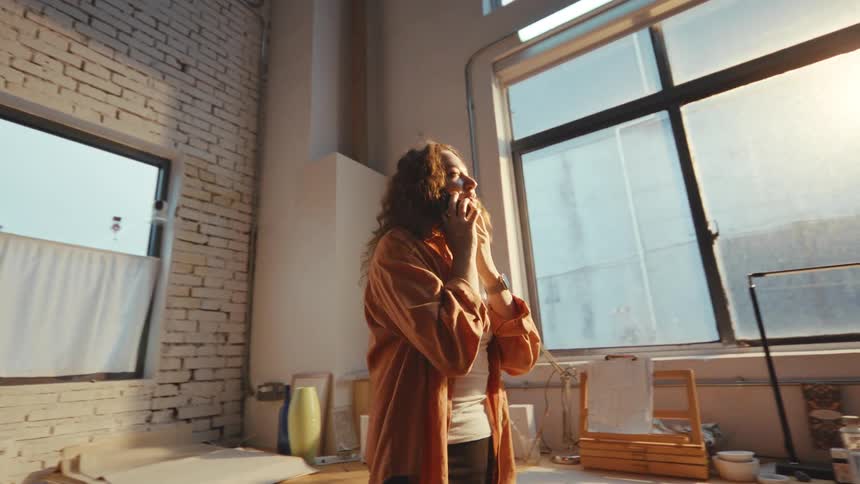 A happy young woman with curly hair talks on her phone while standing in a sunlit industrial loft studio