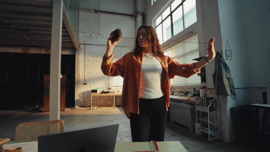 A creative woman stands in her spacious, sun-drenched workshop, slowly and thoughtfully pulling a thread from a ball of yarn