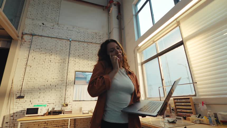 A woman multitasks by talking on her phone while holding and typing on a laptop in a sunlit industrial workshop