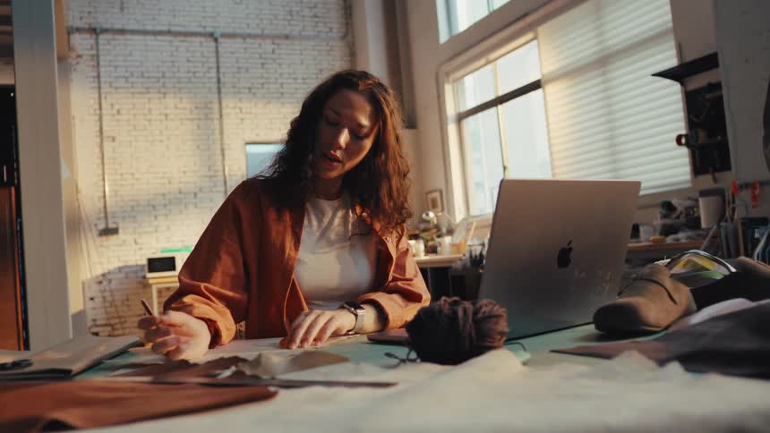A young female artisan sketches a new design pattern for leather goods at her desk in a sunlit workshop