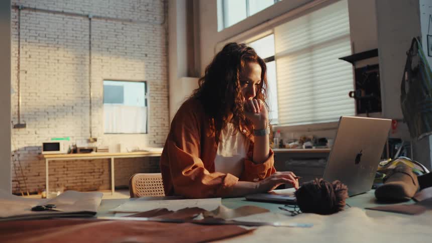 A young female artisan sketches a new design pattern for leather goods at her desk in a sunlit workshop