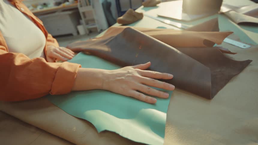 A female artisan carefully examines and touches various colorful pieces of genuine leather in her sunlit workshop