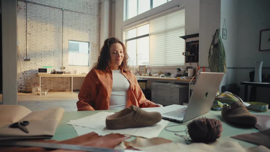 A young female shoe designer finishes her work on a laptop and relaxes, feeling a sense of accomplishment in her sunlit studio