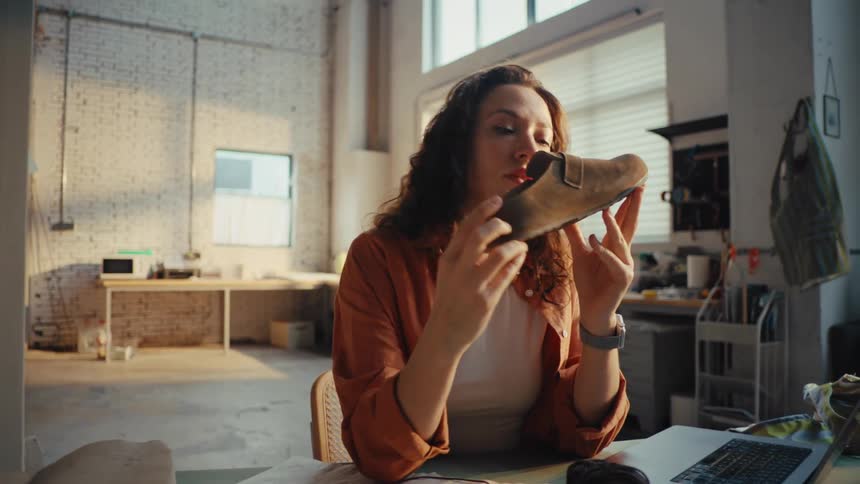 A young female shoemaker carefully inspects and smells a newly handcrafted suede shoe in her sunlit workshop