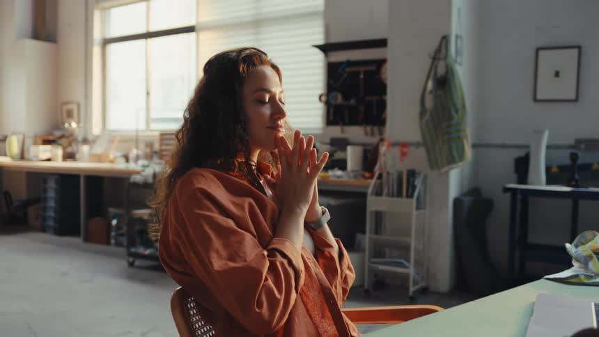 A female artisan works diligently on her laptop in a sunlit studio, then leans back in her chair to take a calm, reflective break