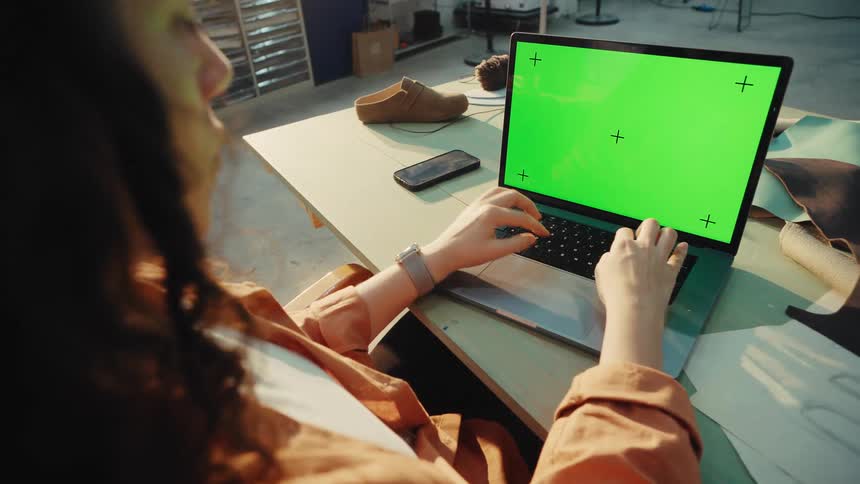 Over-the-shoulder shot of a female designer working on her laptop with a green screen in her sunlit workshop