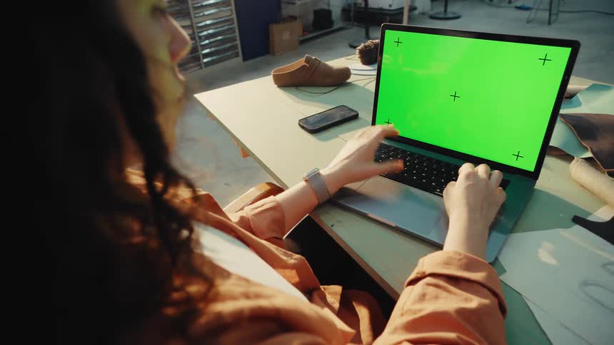 Over-the-shoulder shot of a female designer working on her laptop with a green screen in her sunlit workshop